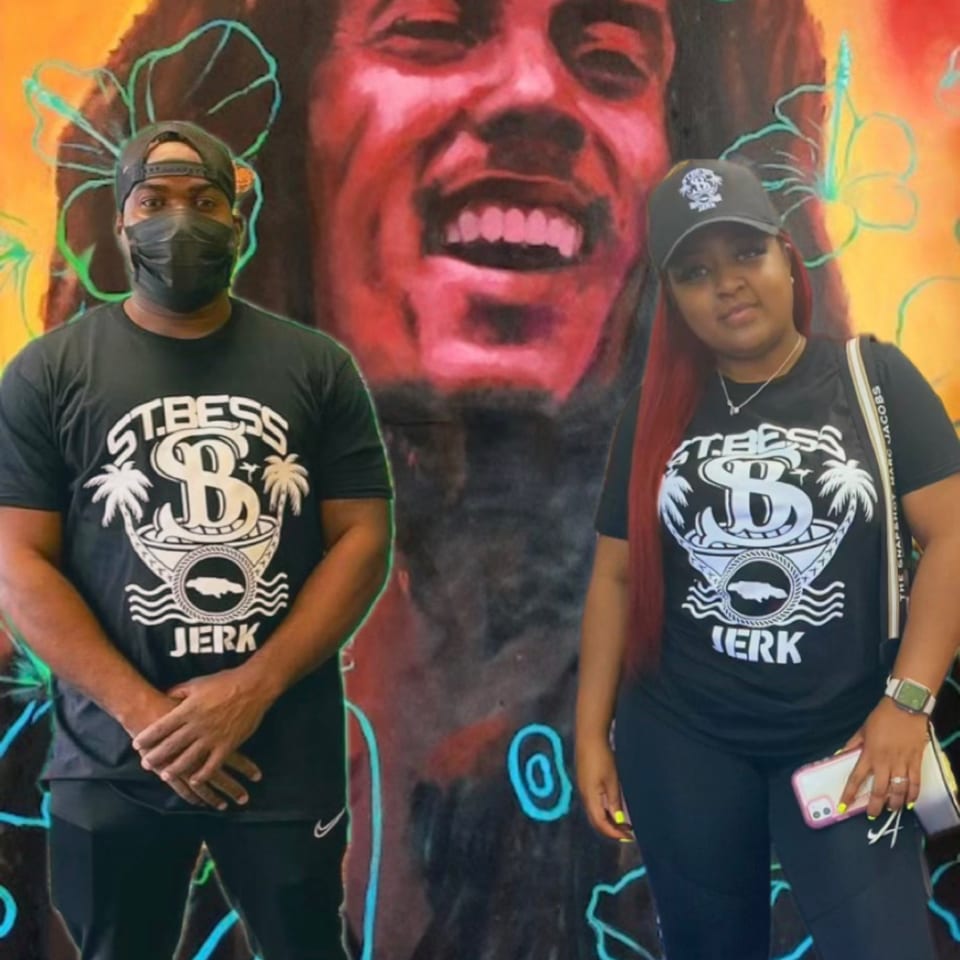 Two people in St. Bess Jerk shirts stand before a colorful mural of Bob Marley.