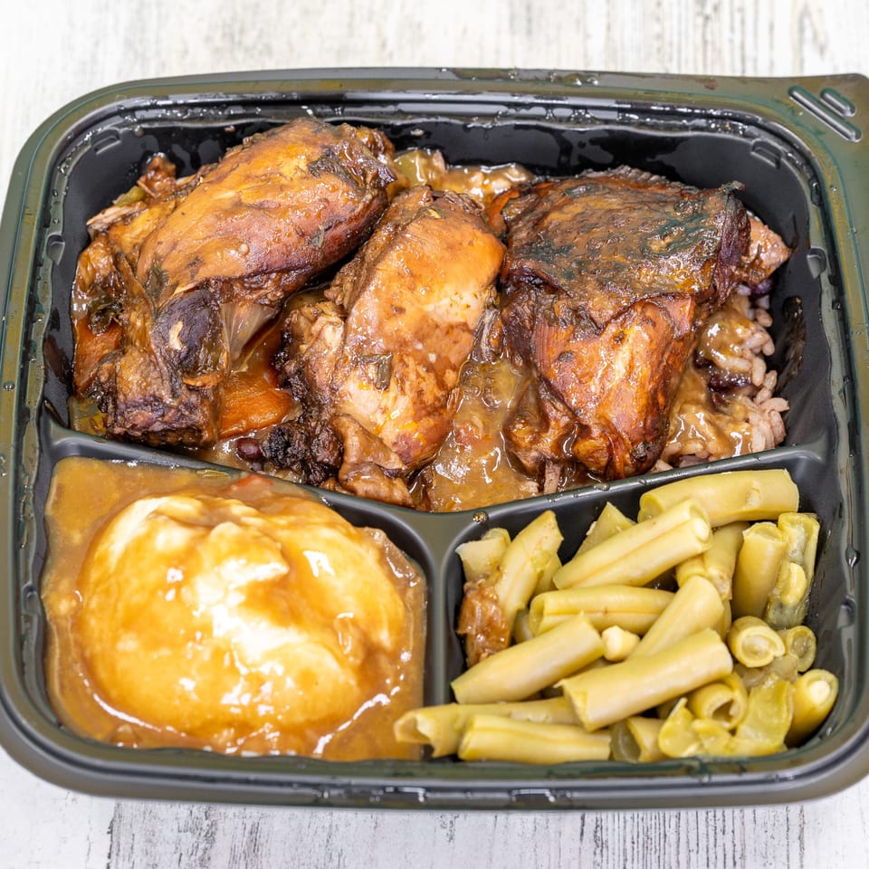 St. Bess Jerk meal with brown stew chicken, rice, mashed potatoes, and green beans in a black container.