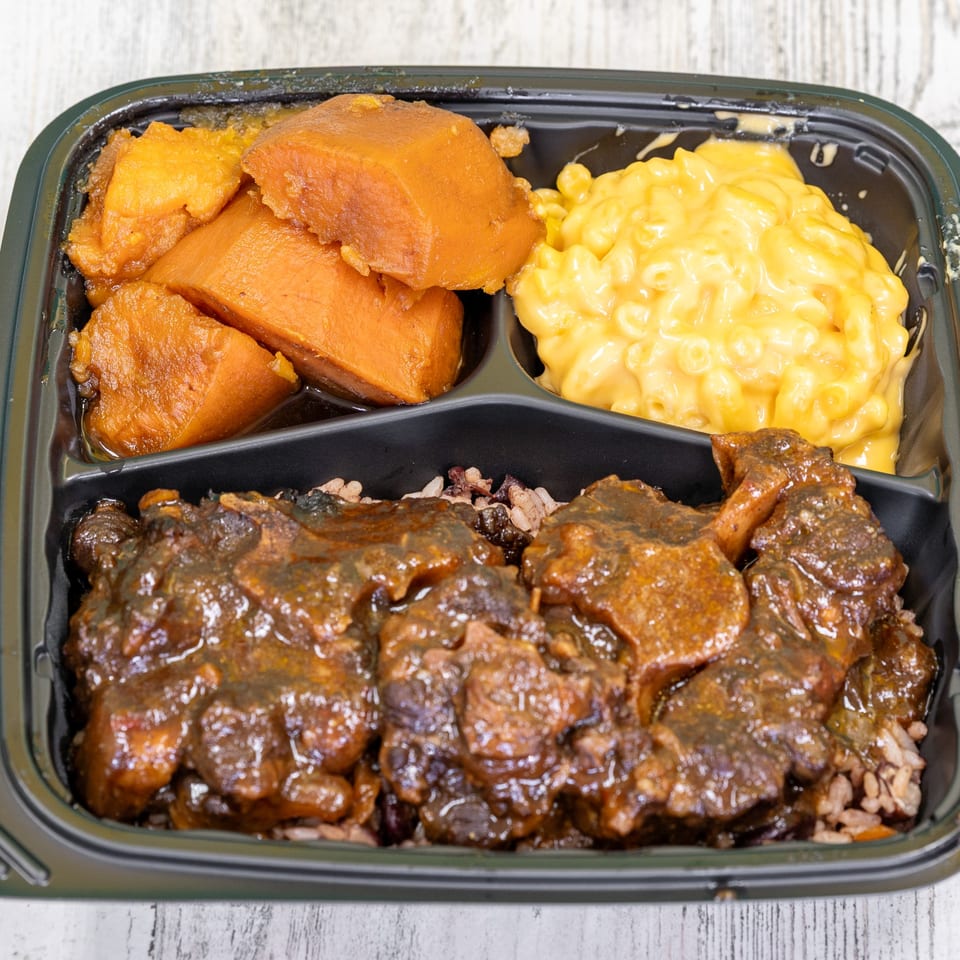 Jamaican oxtail stew with rice and peas, mac and cheese, and candied yams in a black takeout container.