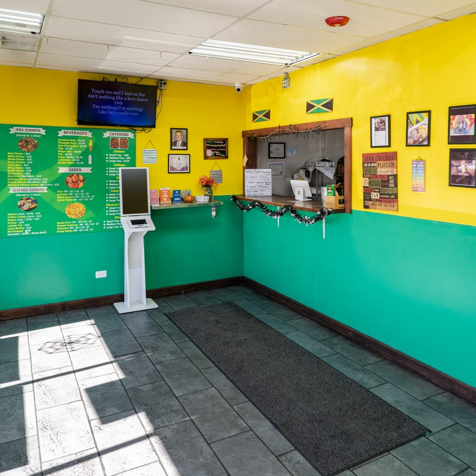 The interior of St. Bess Jerk features bright yellow and teal walls, a menu board, and a self-order kiosk.
